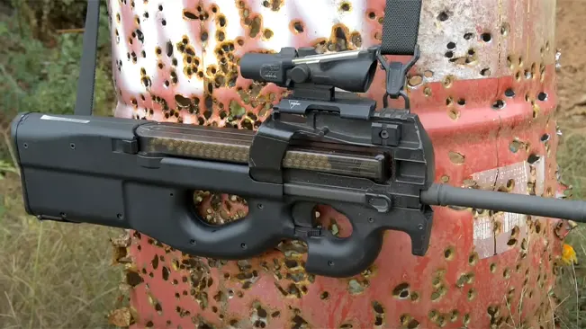 FN PS90 with mounted sight leaning against a bullet-riddled rusted barrel