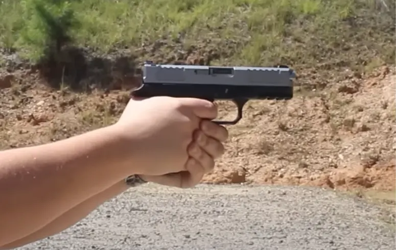 A person's hands holding a black handgun aimed forward, with a natural, earthy background.