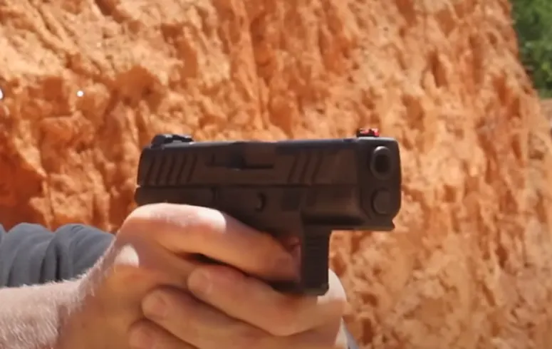 Hands gripping a black handgun with a red-dot sight, set against a red clay background.