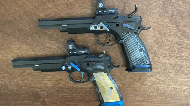 Two competition pistols with optical sights, one with a blue and gold grip and the other with a black and silver checkered grip, both lying on a wooden surface.