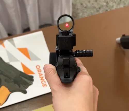 First-person view of a person aiming a pistol with a red dot sight, with the focus on the sight's reticle and a blurred target in the background.