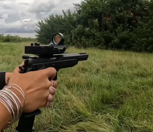 A shooter's hands holding a CZ 75 TS Czechmate with a mounted red dot sight, aiming at a target in a grassy field with trees in the background.