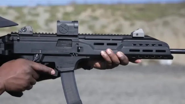 Hand holding a CZ Scorpion Evo 3 S1 carbine with red dot sight and angled foregrip.