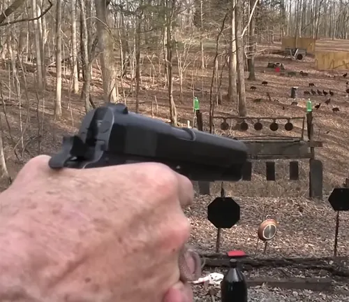 Close-up of a person's hand firing a Colt 1911 pistol at an outdoor shooting range with targets in the background