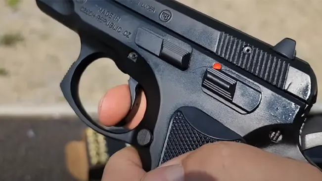 Close-up view of a person's hand with a finger on the trigger of a CZ 75 B 9mm pistol, the safety is on as indicated by the red dot.