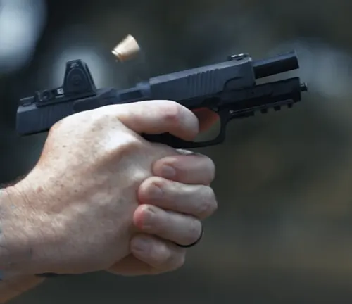 Close-up of a person's hand firing a semi-automatic pistol with a mounted reflex sight, ejecting a brass casing mid-flight against a blurred background.