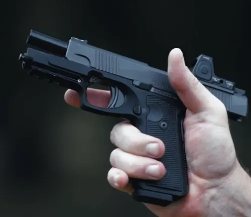 A person's hand holding a black semi-automatic pistol with an attached reflex sight, against a blurred background.