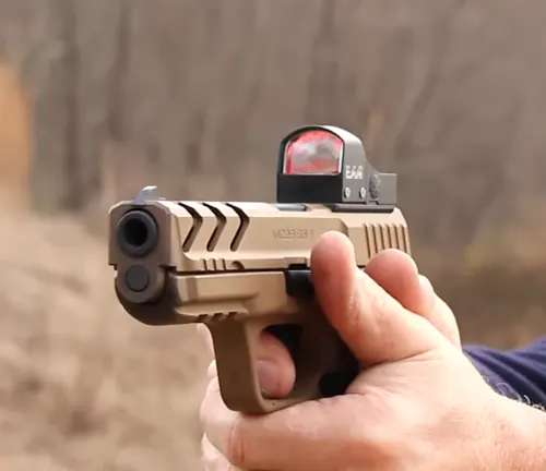 A person holding a tan and black EAA Girsan MC-28 pistol with a mounted red dot sight, ready to fire, with a blurred natural background indicating an outdoor setting.