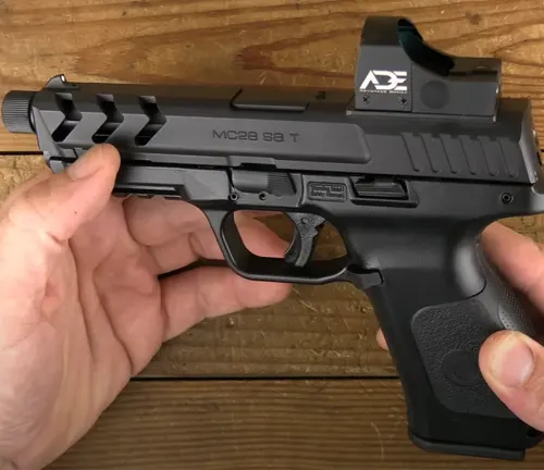 A hand holding a black EAA Girsan MC28 SA-T pistol with a mounted reflex sight, showcasing its slide cuts and textured grip against a wooden background.