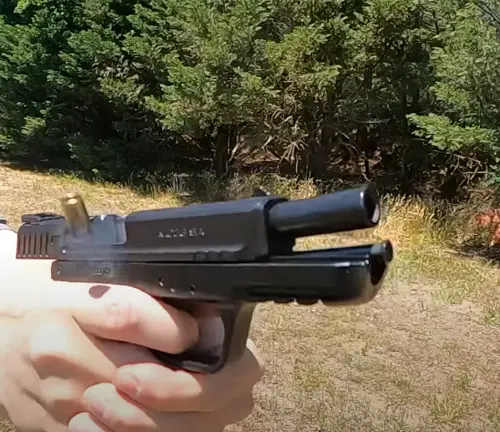 A hand holding a black EAA Girsan MC28 SA pistol with a brass ejection in progress, set against a backdrop of greenery and outdoor terrain.