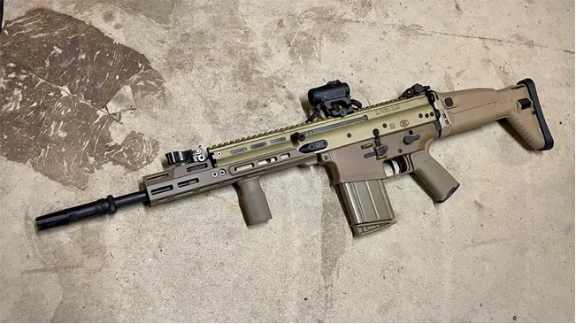 Desert tan FN SCAR 17S rifle with a red dot optic sight on a concrete floor