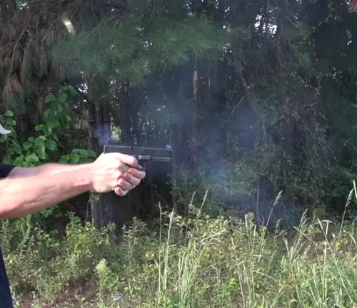 An individual firing a Glock 21 semi-automatic pistol outdoors, with visible muzzle flash and smoke against a backdrop of greenery.