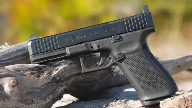 A Glock pistol resting on a gnarled piece of wood with a blurred natural background, highlighting the firearm's slide markings and profile.