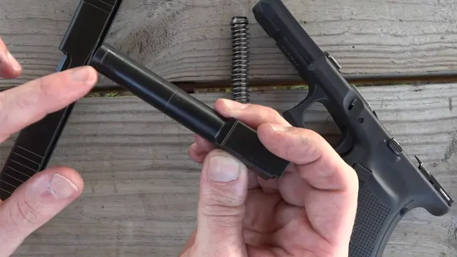 A person's hands performing field stripping of a Glock pistol, with the barrel and recoil spring assembly being removed from the slide, on a wooden background.