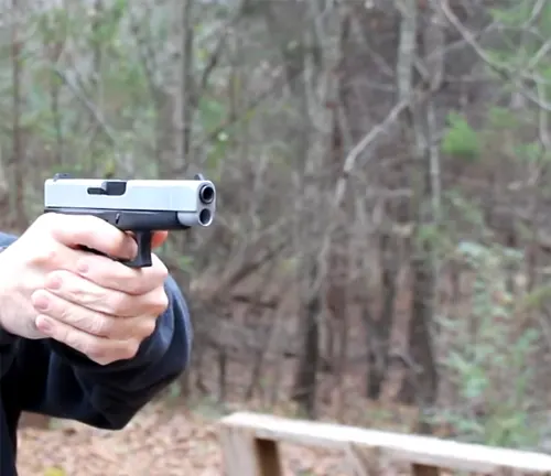 Person holding a two-tone Glock 48 pistol with a silver slide, aimed downrange with a blurred natural backdrop.