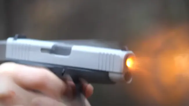 A close-up of a two-tone Glock 48 pistol being fired, with a visible muzzle flash from the barrel against a blurred background.