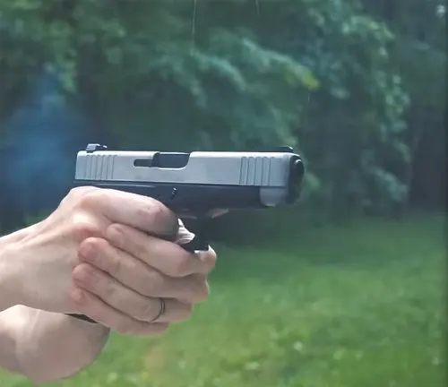 Hands holding a Glock 48 pistol with a silver slide, aimed and ready to fire, with a green blurry background indicating outdoor conditions.