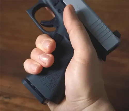 Close-up of a person's hand with a strong grip on the textured handle of a Glock 48 pistol with a silver slide, against a wooden floor background.