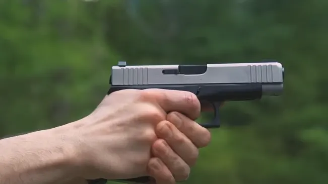 A person's hands holding a Glock 48 pistol with a silver slide, aiming steadily against a blurred green foliage background.