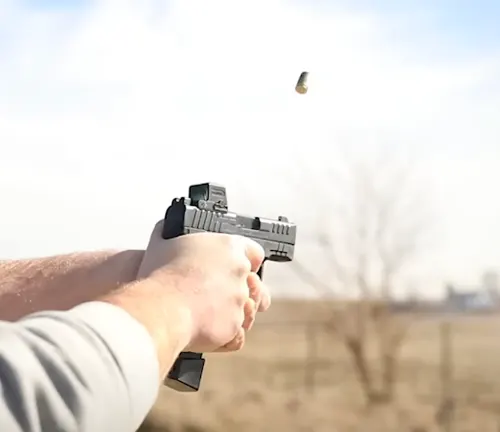 A shooter's hands gripping an IWI Masada Slim pistol mid-firing, with a spent casing ejecting from the firearm, against a blurred outdoor backdrop.