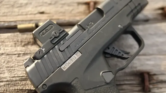 Close-up of an IWI Masada Slim pistol lying on a wooden surface, with a Holosun red dot sight mounted on the slide and a spent bullet casing beside it.