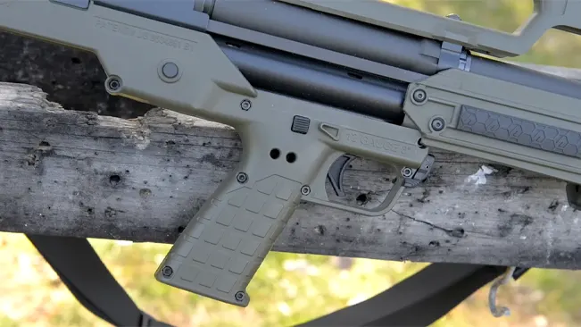 Close-up of the grip and trigger area of an olive drab Kel-Tec KS7 shotgun, mounted on a wooden beam.