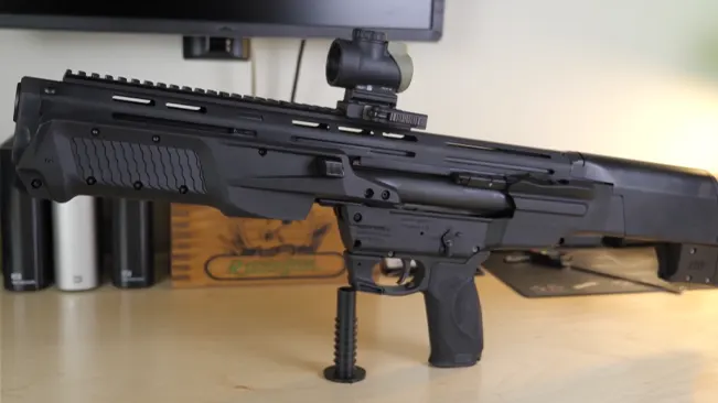 A Smith & Wesson M&P 12 shotgun is displayed on a stand, featuring a mounted optical scope and bipod support, against an indoor background.