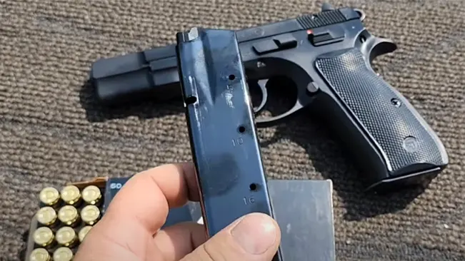 A person's hand holding an empty magazine in front of a CZ 75 B handgun with a box of bullets and a magazine loader in the background.
