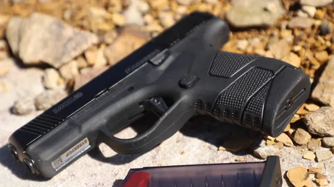 A Mossberg MC1SC pistol lying on a rocky surface with its magazine removed, visible in the foreground, highlighting the pistol's textured grip and trigger guard.