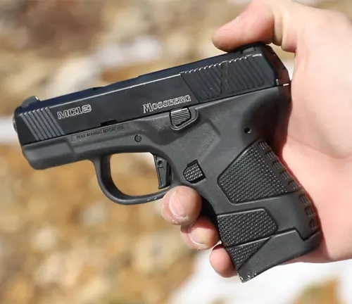 A hand holding a Mossberg MC1SC pistol, displaying its compact size, black polymer frame, and textured grip against a blurred outdoor background.