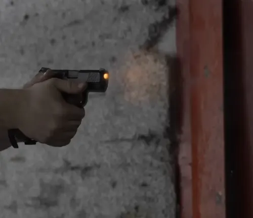 A hand firing a Mossberg MC1SC pistol with a visible muzzle flash, in a blurred indoor range setting.