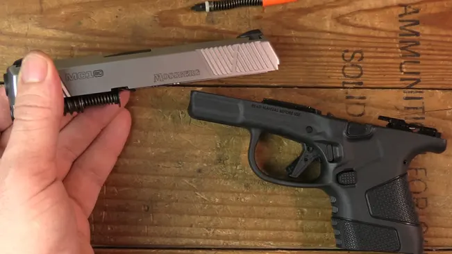 A hand holding the disassembled slide and recoil spring of a Mossberg MC1SC, with the frame and other maintenance tools visible on a wooden bench.