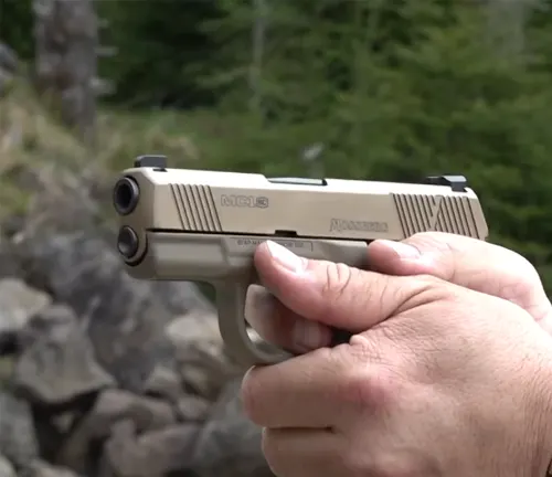 A hand holding a two-tone Mossberg MC1SC pistol with a stainless steel slide, aimed forward with a natural, rocky background.