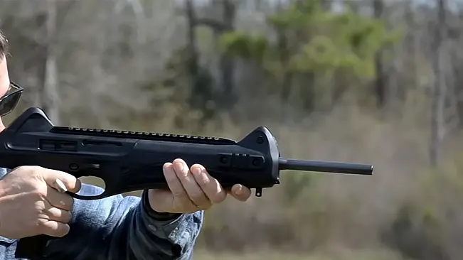 Person holding a modern firearm with a barrel shroud and tactical rail, outdoors.