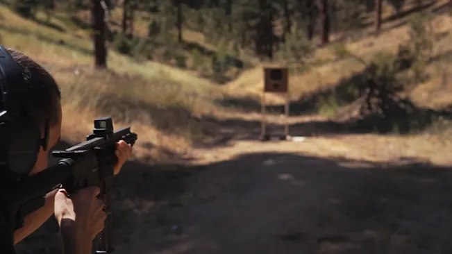 A shooter with ear protection is aiming a rifle with a mounted optic at a target downrange in a forested area. The focus is on the shooter, making the background target slightly blurred.