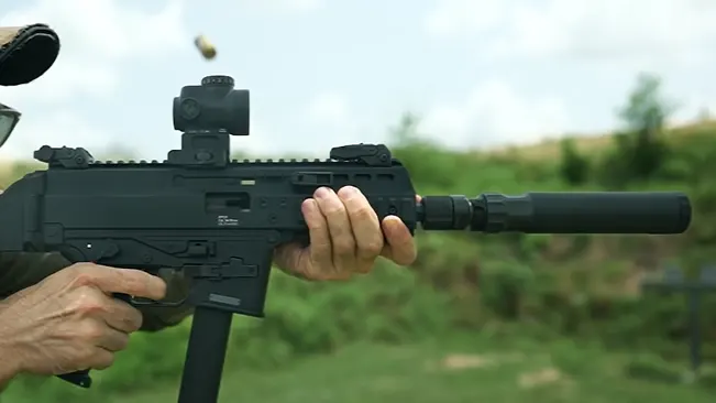 A person is holding a black tactical rifle with a suppressor and a red dot sight. The rifle is mid-firing with a casing being ejected, set against a natural, green background.