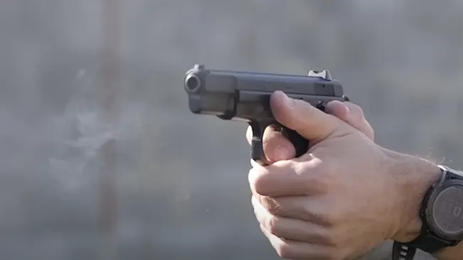 A close-up of a handgun being fired, with smoke visible near the barrel, held by a person's hands with a wristwatch visible on the left hand.