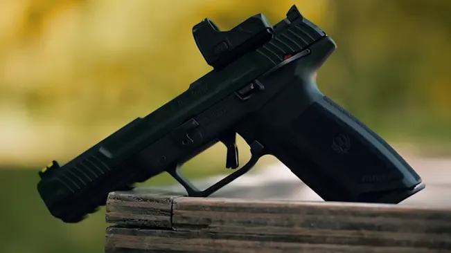 A Ruger 5.7 pistol with an optic attachment, resting vertically on a weathered wooden plank, set against a soft-focus background of yellow and green foliage.