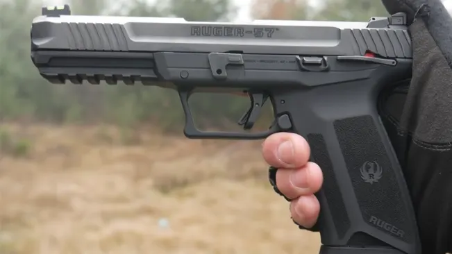 Close-up of a hand gripping a Ruger 5.7 pistol, with clear view of the textured grip and slide, against a blurred outdoor background.