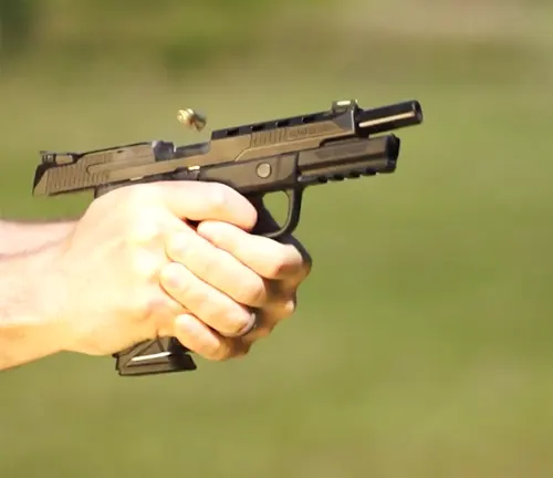 Close-up of a Ruger American Competition pistol being fired, with the slide in rearward motion and a spent casing being ejected, held in a right-handed grip against a blurred natural background.