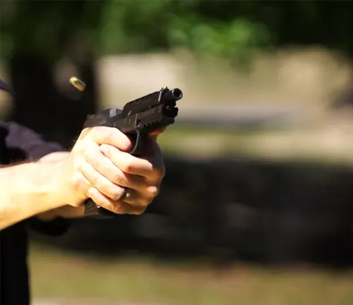 A person's hands firmly gripping a Ruger American Competition pistol during firing, with the muzzle flash visible and a spent casing in mid-ejection, set against a blurred outdoor backdrop.