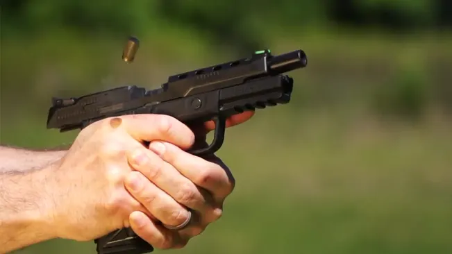 Close-up of a Ruger American Competition pistol being fired, with the slide in rearward motion and a spent casing ejected, held with a two-handed grip against a green outdoor background.