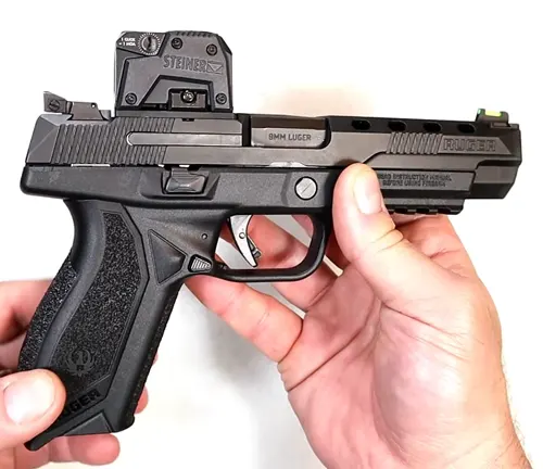 Hands holding a Ruger American Competition pistol with a mounted Steiner red dot sight, showcasing the firearm's black frame, slide serrations, and the caliber marking of 9mm Luger.