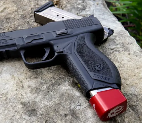 A Ruger American Competition pistol lying on a rough stone surface with a red magazine well extension and a stainless steel magazine beside it, showcasing its ergonomic grip and slide design.