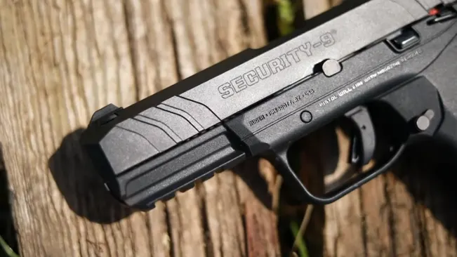 Close-up of the upper slide of a Ruger Security-9 pistol, highlighting the 'SECURITY-9' engraving, front sight, and slide serrations, set against a wooden background.