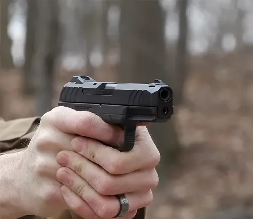 A person with a ring on their finger is aiming a Ruger Security-9 pistol, focused on the front view showing the barrel and front sight, with a blurred forest background.