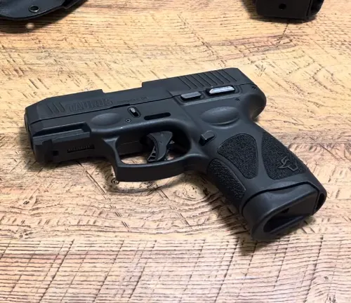 A Taurus G3C semi-automatic pistol with a black polymer frame and matte black steel slide is lying on a wooden surface, with a textured grip clearly visible and the magazine removed, placed beside the firearm.