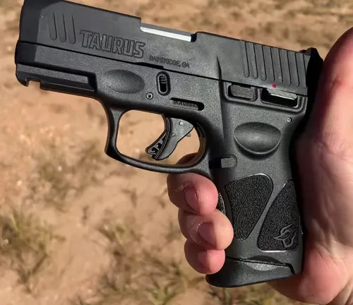 Close-up of a Taurus G3C pistol being held in a person's hand, highlighting the detailed texture of the grip, the trigger guard, and the slide with visible brand markings, against an outdoor, sandy backdrop.