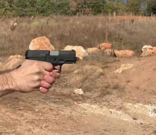 A person's hand is firing a Taurus G3C pistol with a spent casing ejecting from the gun, set against a natural outdoor range with rocks and sparse vegetation in the background.