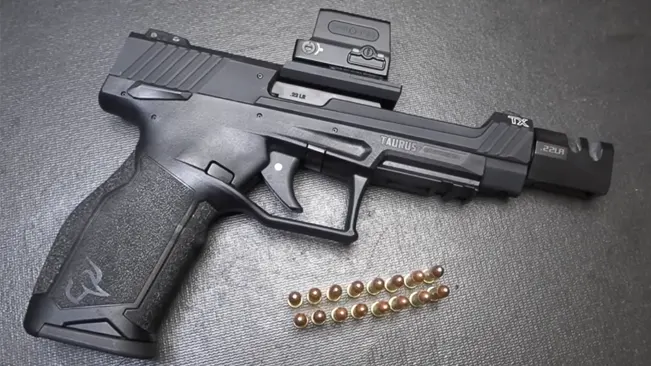 A Taurus TX22 Competition SCR pistol with a mounted red dot sight lying on a textured surface next to a row of .22 LR cartridges.